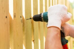 Hands of the carpenter holding electric screwdriver up to wooden fence post